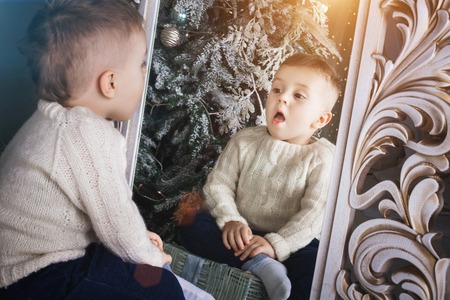 surprised little boy wearing knitted sweater sitting in front of the tracery mirror near the christmas treeの写真素材