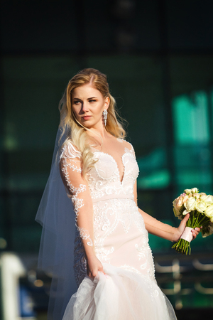 beautiful bride holding a skirt of her wedding dress and bouquet standing on stairs of modern building. wind playing with her blonde hairの写真素材