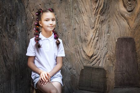 pretty lovely young girl with pigtails wearing white shirt sitting on the chair made from stump near the stem of the tree in the parkの写真素材