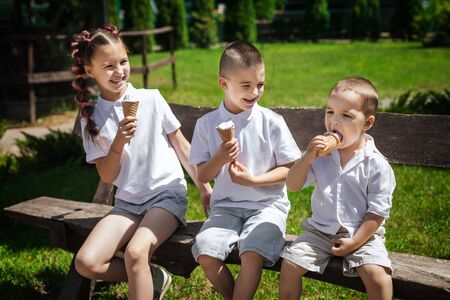 adorable cute brothers and a sister eating ice cream sitting on a wooden bench in the park. fun holiday conceptの写真素材