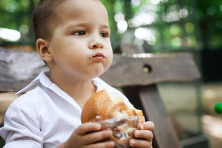 adorable toddler boy eating a bun sitting on a wooden bench in the park. fun holiday conceptの写真素材