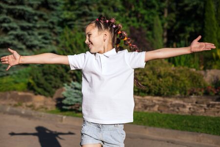 joyful child girl with pigtails waving on the wind wearing white shirt running spreading hands in a sunny summer park. pine trees on a backgroundの写真素材