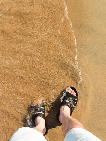 A sea wave comes to my feet when I stand in the sand in black slippers and wait. Copy space. Top down viewの写真素材