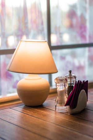 A lamp with a shade on the table in front of the window. A table in a cafe with spices and napkins. Cozy atmosphere in the cafeteriaの写真素材