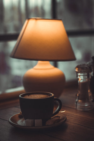 Orange lamp and cup of coffee on the table. Cozy atmosphere, dim lights, evening in a cafe. A cup of coffee on a platter against the background of a lamp with a lampshade. Atmospheric dark photo on the theme of coffeeの写真素材