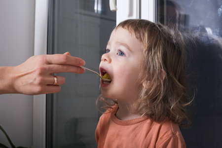 A little girl is eating from a spoon in the kitchen at homeの写真素材