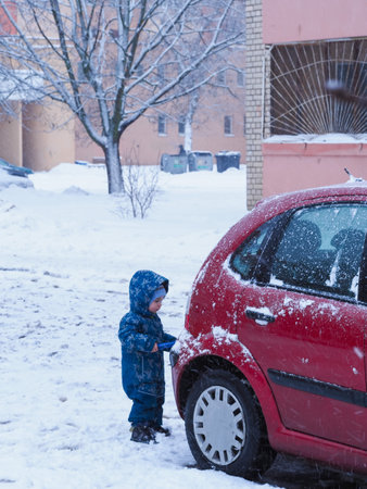 Little boy in winter clothes stands near the car and looks at the snowの写真素材