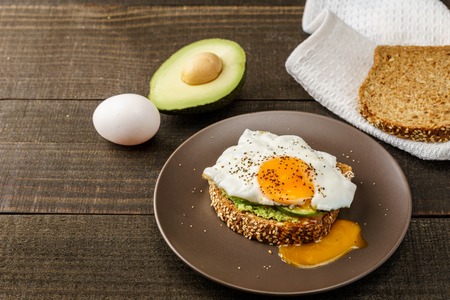 sandwich with avocado and a fried egg on a brown plate on a rustic wood and table bread on white kitchen towelの写真素材
