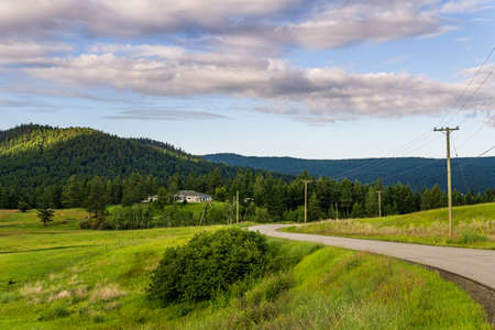 KAMLOOPS, CANADA - JULY 10, 2020: country road through the medow landscape with house on background and cloudy skyのeditorial素材