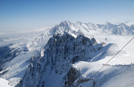 White steep peaks of the Alps against blue sky and snow slope with mountain-skiers, Mont Blanc, steel rope in the foregroundの写真素材