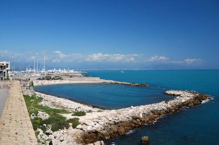 Aquamarine sea and azure sky of mediterranean settlement Antibes, pier with vessels and yachts in the background, beach fenced with stone foregroundの写真素材