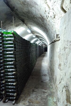 Long piles of wine bottles are in the stone wine-cellar located at the foot of a mountain. Narrow...