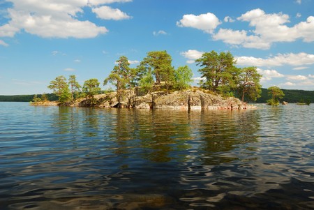 Small island is in the middle of clear large lake. In the foreground there is waving surface of transparent water. Some green trees grow on stony ground under the summer sky.の写真素材