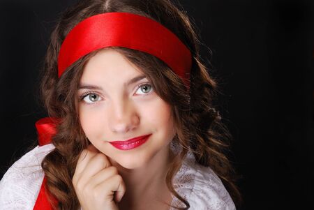 Pretty teenage girl is looking at the camera friendly. Her dark long hair is tied with a red ribbon. She is photographed in dark background.の写真素材