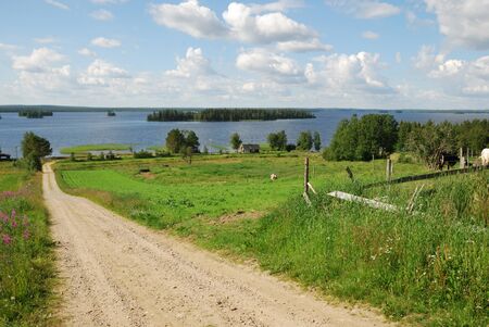 Country natural road is running between green fields and pastures.  In the background there are blue water of big forest lake and the summer sky with clouds.の写真素材