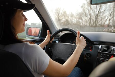 Young woman is holding a steering wheel  She is photographed inside of her car  Happy driver is...