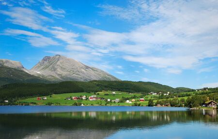 The blue sky and mountains are reflecting in smooth surface of the fjord. In the background a...