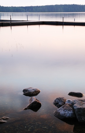 Smooth surface of quiet water reflect the sky at sunset. In the background there are dark silhouettes of a wooden foot bridge and several stones in the foreground.の写真素材