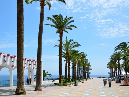 The square is in front of the Balcony of Europe in sunny day. There are a white stone portico with flowers, high palm trees and persons walking in Nerja on the Costa del Sol.のeditorial素材
