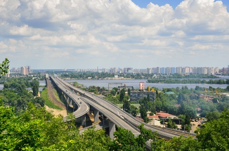 Wide river is dividing the city into right and left riverside  Both banks are overgrown with green trees and bushes  A modern bridge recedes into the left-bank residential district and the cloudy sky の写真素材
