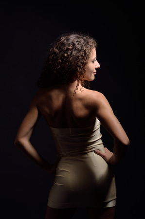 Young curly woman is posing backwards  She is wearing a strapless sheath short dress  Her sexy body is photographed in the dark with few highlights  の写真素材