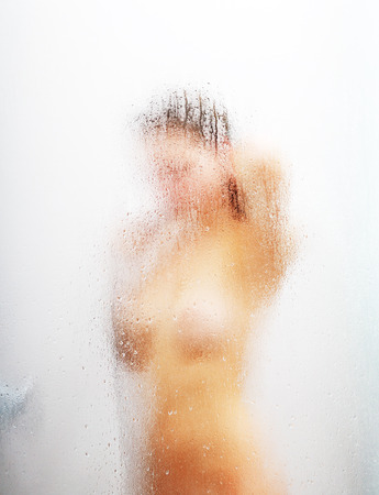 A nude girl is washing in the shower bath. Her blurred silhouette is visible through the sweat glass surface with water drops and streams. Focus is on the wet glass wall of the shower cabin.の写真素材