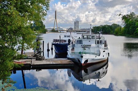 Two boats are moored at the small pier in the Baltic city Riga. The blue sky with white clouds are reflected in the water surface.のeditorial素材