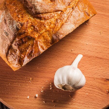 garlic and a half loaf of bread on a wooden board with crumbs looks very appetizing all fresh and...