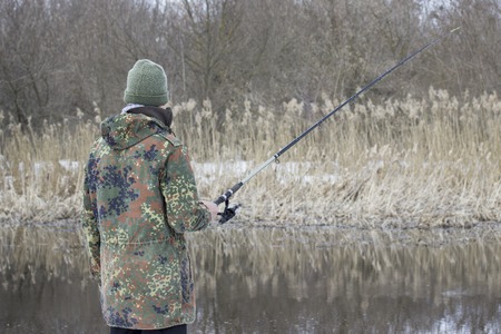 fisherman in camouflage with a spinning in his hand on the background of the river and reeds concept of fishingの写真素材