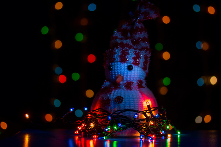 toy snowman standing in garlands on a blurred background of bright multi-colored lights concept of Christmas evening and new yearの写真素材