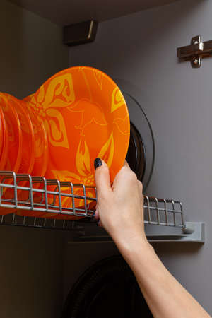 a woman's hand picks up an orange plate from a high-rise kitchen cabinet. High quality photo