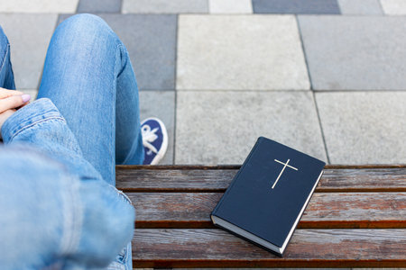 the bible lies on the bench next to the woman. a closed bible lies on a bench next to a woman