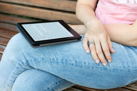 woman reading an e-book sitting on a park bench. reading a book built on electronic ink technology....