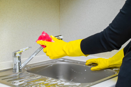 hand in a yellow glove washes a kitchen sink with a sponge. a woman washes a sink in the kitchen, shot close-up.の写真素材