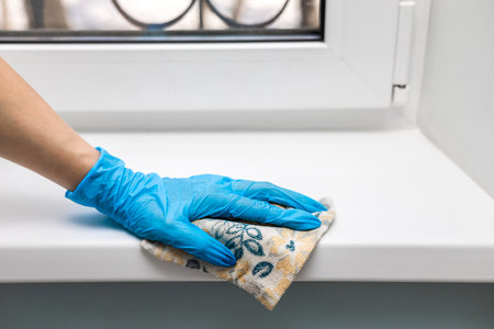 housewife wearing gloves wipes a plastic window sill. a hand in a blue glove wipes the windowsill...