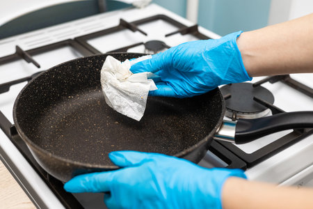 a hand in a rubber glove wipes a frying pan with a paper towel. hand wipes a frying pan in the...