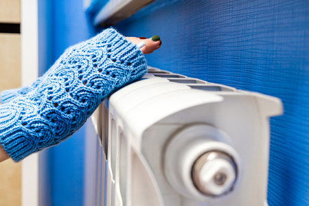 girl in knitted mittens warms her hands on a radiator. girl in knitted fingerless gloves warms her hands on a radiator. woman warming her hands over a radiator. warm your hands at home.の写真素材