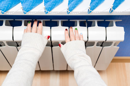 girl in a sweater warms her hands on a radiator. woman warming her hands over a radiator. warm your hands at home. the girl is cold. central heating of the house. a person is cold in winter. female hands near radiatorの写真素材