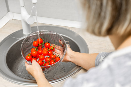 woman washing cherry tomatoes in kitchen sink. woman washing tomatoes in a colander in the kitchen. tomatoes on a branch.の写真素材