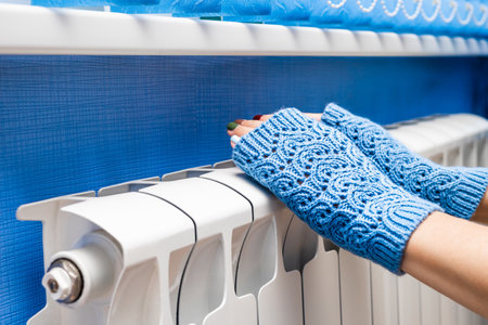 girl in knitted mittens warms her hands on a radiator. girl in knitted fingerless gloves warms her...