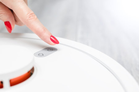 a woman's finger presses the power button of a robot vacuum cleaner. the housewife turns on the...