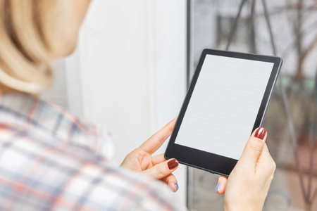 woman reading e-book standing near window in house. girl reading e-book at home. female hand holding...
