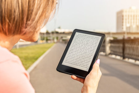 woman reading e-book sitting on bench in city park. woman reading a book sitting on the embankment. ebook mockup.の写真素材