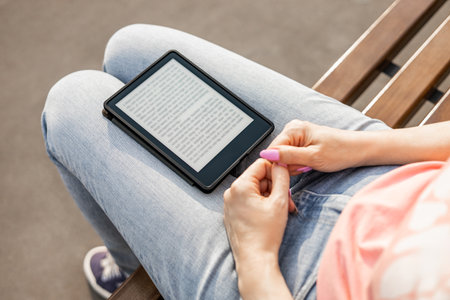 woman reading e-book sitting on bench, close-up. an e-book lies on a girl's lap. ebook mockup.の写真素材