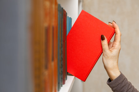 a woman's hand takes a red book from the shelf. A woman in a library takes a book from a bookshelf....