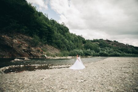 Beautiful luxury young bride in long white wedding dress and veil standing near river with mountains on background.の写真素材