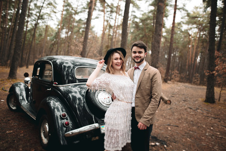 Gorgeous newlywed bride and groom posing in pine forest near retro car in their wedding day.