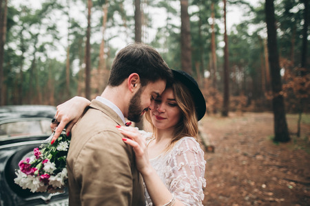 Gorgeous newlywed bride and groom posing in pine forest near retro car in their wedding day.の写真素材
