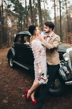 Gorgeous newlywed bride and groom posing in pine forest near retro car in their wedding day.の写真素材