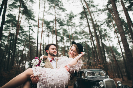 Gorgeous newlywed bride and groom posing in pine forest near retro car in their wedding day.の写真素材